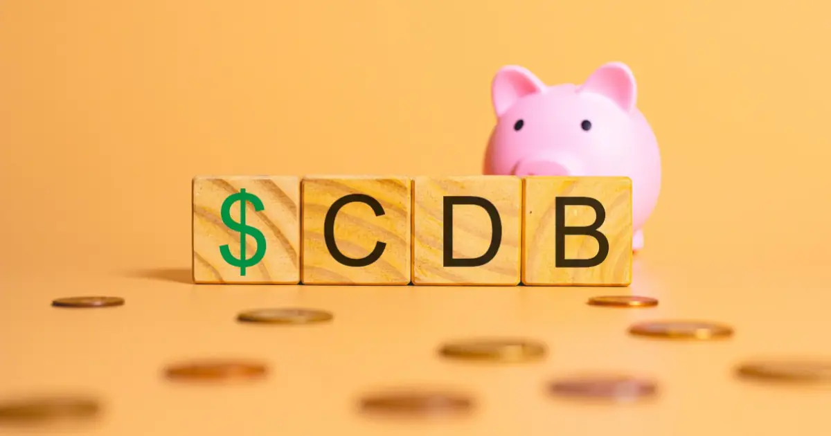 Blocos de madeira formando a sigla CDB, precedidos por um cifrão verde, ao lado de um cofre-porquinho rosa, com moedas espalhadas sobre fundo alaranjado